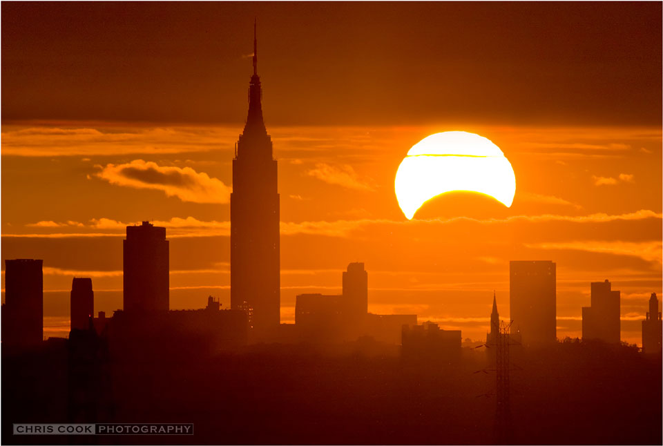 Über der Silhouette von New York geht die Sonne als angeschnittene Scheibe auf. Ein Teil der Sonne war vom Mond bedeckt. Am orange gefärbten Himmel wirkt die Finsternis sehr dramatisch.