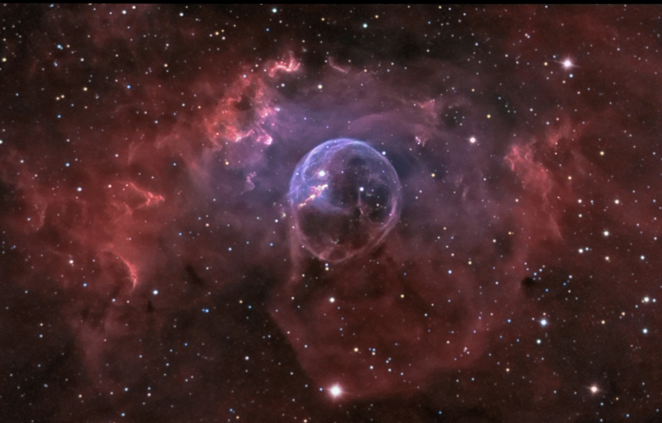 Umgeben fon einem Kranz schwach leuchtender kaminroter Wolken leuchtet eine zarte Blase in der Mitte. Im Bild sind wenige Sterne verteilt.