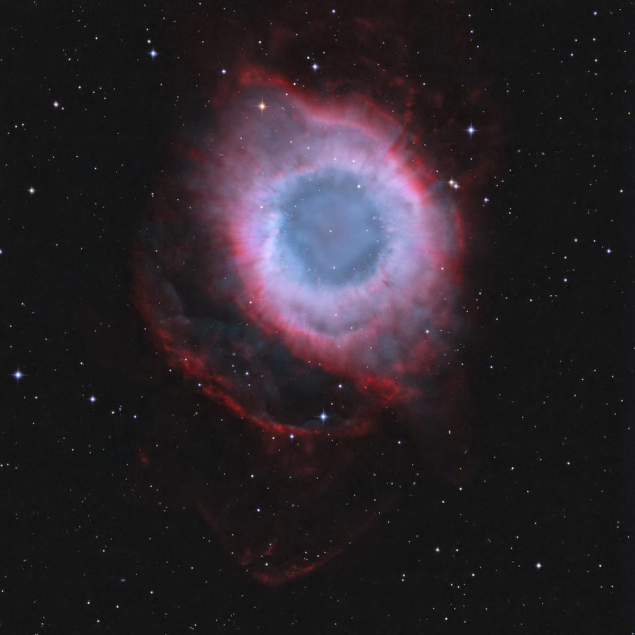 Mitten im Bild leuchtet ein ringförmiger Nebel. In der Mitte ist eine dunkelblaue Scheibe, darum herum ist ein fliederfarbener Nebel mit starken karmesinroten Rändern.