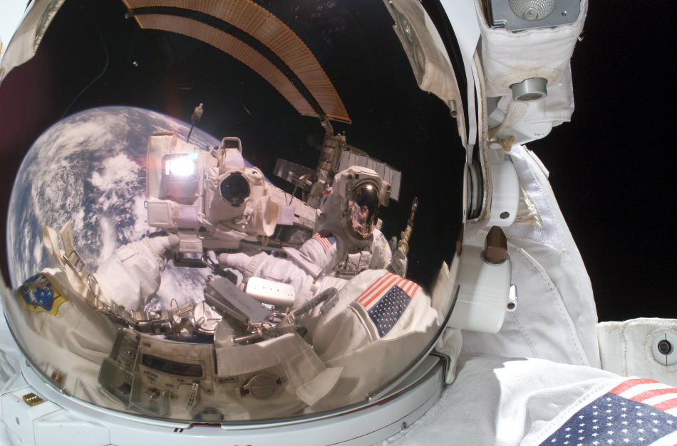 Im Visier eines Astronautenhelms spiegeln sich die Erde, Teile der Internationalen Raumstation ISS und ein zweiter Astronaut.