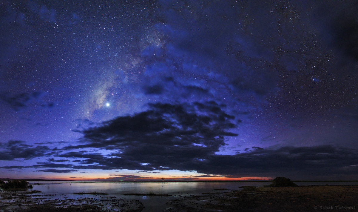 Über dem stillen Turkanasee in Kenia leuchten Sterne und die Milchstraße hinter dunklen Wolken. Links oben gleißt die Venus als Abendstern.