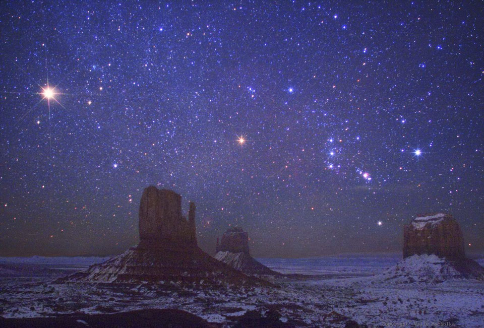 Über dem Monument Valley mit seinen ikonischen Tafelbergen, den Mitten und Merrick Butte, strahlt ein prachtvoller Sternenhimmel. Rechts liegt das Sternbild Orion mit Gürtel, Beteigeuze und Rigel. Links ist das gleißende Licht des Planeten Mars.