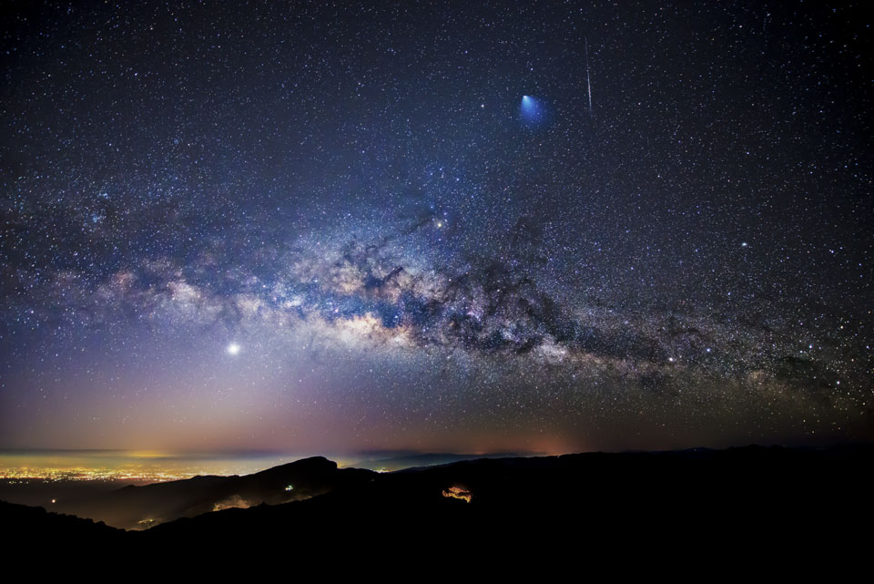 An einem surreal wirkenden Sternenhimmel schwebt das Band der Milchstraße. Links leuchtet der Planet Venus, unten schimmern die Lichter von Städten, oben rechts zischt ein Meteor, daneben ist die blau leuchtende Abgasfahne einer startenden Ariane 5.