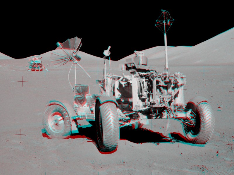 Das graue Bild hat rote und cyanfarbene Ränder. Es ist ein Anaglyphenbild und wirkt dreidimensional, wenn man es mit rot-blauen Brillen betrachtet. Vorne steht ein großer Mondrover, hinten sind Mondberge und das Mondlandemodul der Mission Apollo 17.
