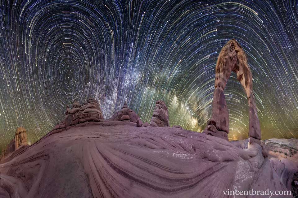 Hinter geschichteten Sandsteingebilden ziehen Strichspuren von Sternen in gekrümmten Bögen über den Himmel. In der Bildmitte wölbt sich die Milchstraße. Das Bild ist ein 360-Grad-Panorama, das waagrecht komprimiert wurde.