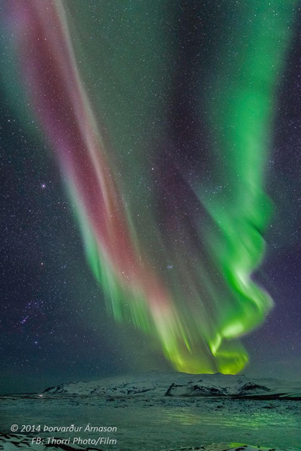 Über einem verschneiten Stratovulkan, dem Öræfajökull auf Island, strahlt eine leuchtend grüne Polarlichtschau mit einigen purpurfarbenen Strähnen. Der Himmel ist dicht von einem Sternenteppich bedeckt. Unten im Schleier leuchten die Plejaden und die Hyaden, links ist das Sternbild Orion.