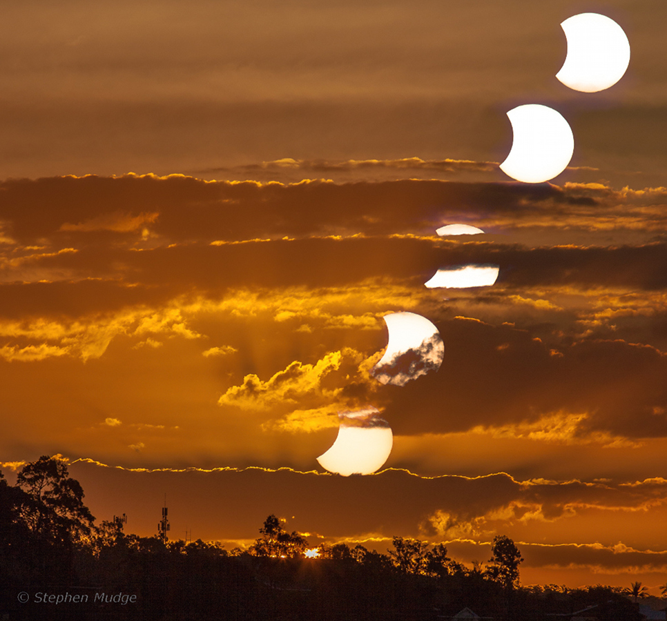 Am dramatisch orange gefärbten, stark bewölkten Himmel bei Brisbane in Australien geht die Sonne unter. Sie ist teilweise vom Mond bedeckt.