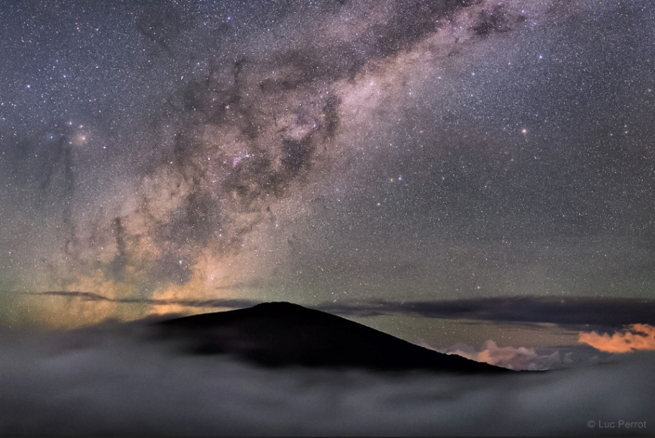 Über einem Vulkangipfel auf der französischen Insel La Réunion geht die Milchstraße mit ihren dunklen Wolken auf. Der Gipfel ragt aus einem Wolkenmeer.