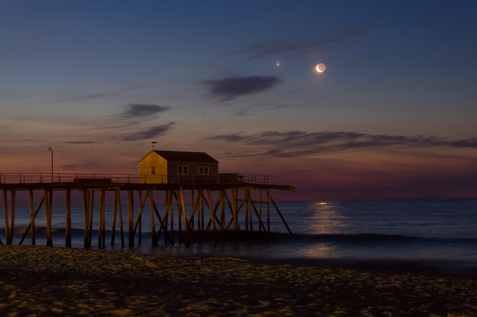 Über dem Meer leuchten Venus und Sichelmond in der Morgendämmerung. Der Himmel ist unten rötlich und oben dunkelblau gefärbt. Am Strand steht ein Holzgebäude auf Stelzen.