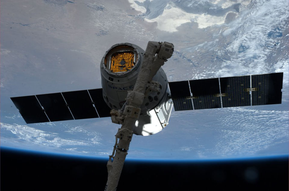 Die Erde füllt fast das ganze Bild, unten ist der Rand zu sehen. Der Blick fällt auf Meere und Wolken, links oben ist eine Landmasse. Vorne fängt der Canadarm2 der ISS gerade eine Dragon-Kapsel ein.