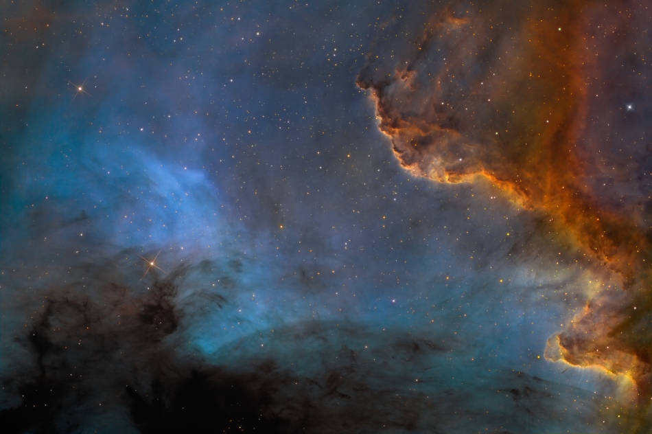 In der Mitte leuchtet ein blauer flächiger Nebel, der links unten von dunklen Wolken verdeckt wird. Rechts und oben ist eine orange-braun-farbene Wolke mit einem klar abgegrenzten Wall am Rand.