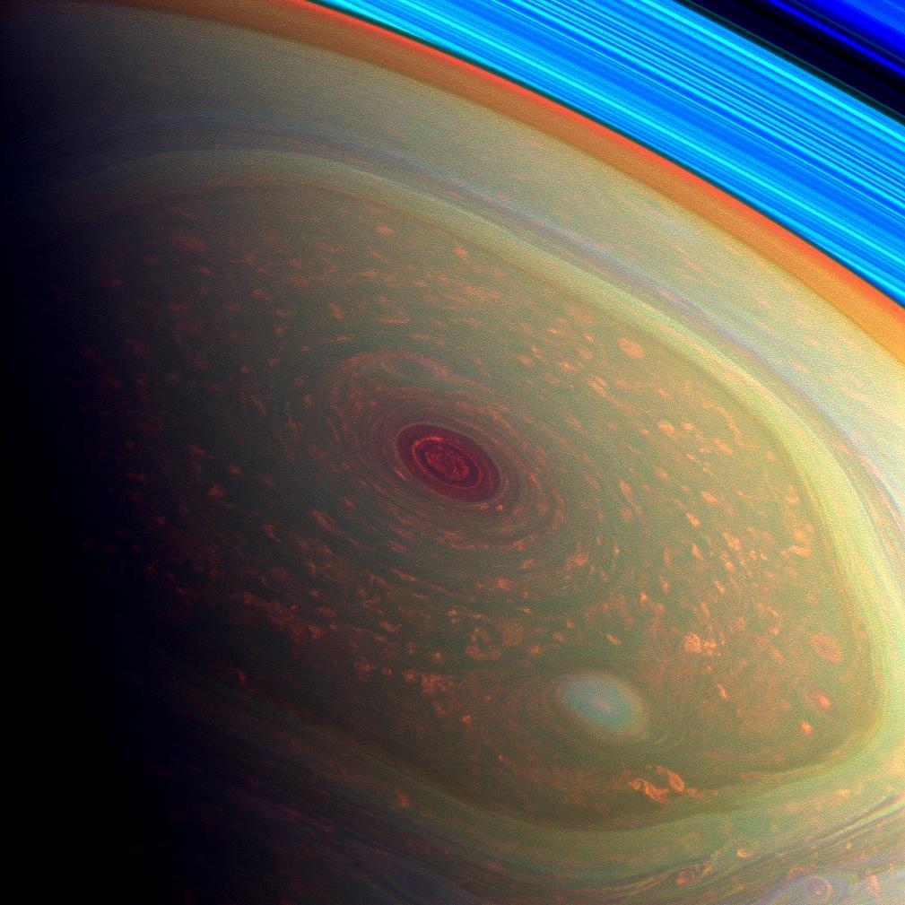 Saturns Nordpol rückte 2012 ins Sonnenlicht. Hier befindet er sich an der Licht-Schatten-Grenze, dem Terminator. In der Mitte ist ein dunkelroter runder Strudel, außen herum verläuft eine sechsseitige Sturmstruktur. Rechts oben leuchten die Saturnringe in hellem Blau.