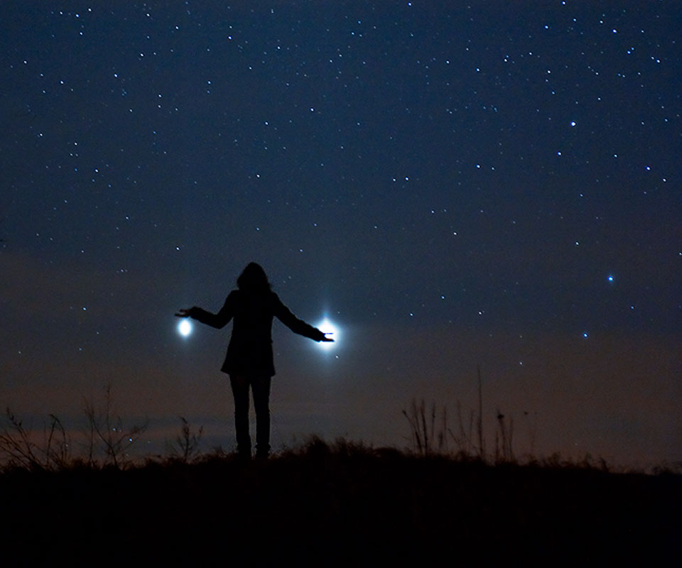 Am Horizont steht eine Person. Sie hält die Planeten Venus und Jupiter scheinbar in den Händen. Am dunkelblauen Himmel leuchten Sterne, am Horizont ist das letzte Abendrot.