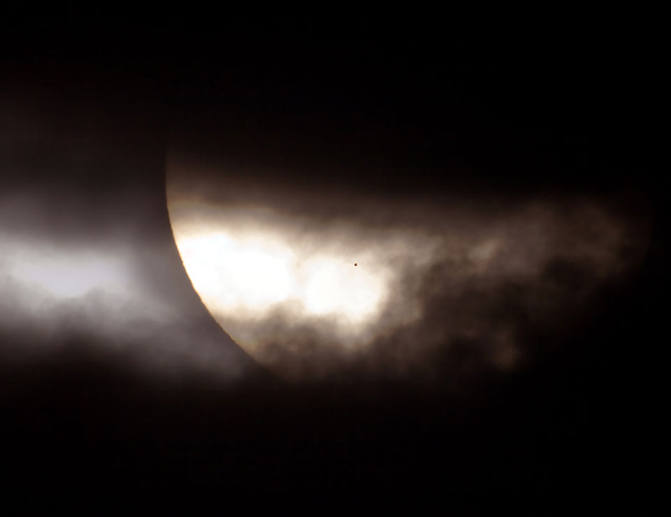 Zwischen sehr dunklen Wolken schaut in der Mitte ein Teil der Sonne hervor, darauf ist ein winziger dunkler Fleck, nämlich der Planet Merkur.