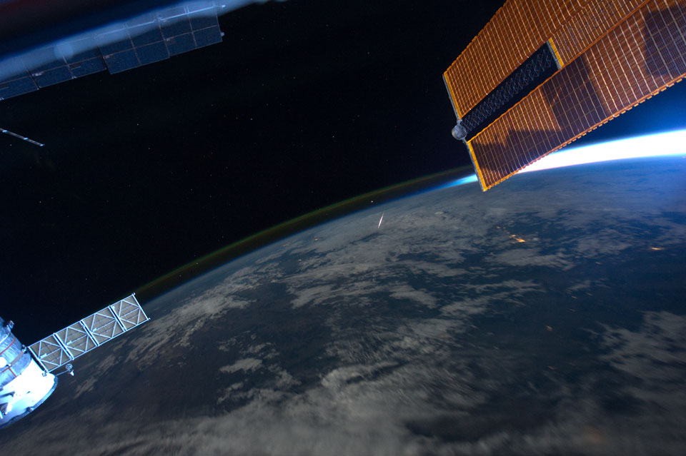 Über der Erde blitzt in der Bildmitte ein Meteor auf. Am Rand der Erde leuchtet grünliches Nachthimmellicht. Links und rechts oben sind Bauteile der ISS zu sehen, rechts unten ist eine Kapsel.