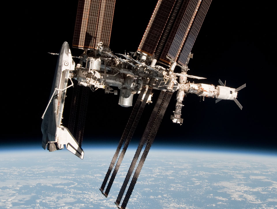 Über der Erde schwebt die Internationale Raumstation mit Solarpaneelen und Modulen. Links ist eine Raumfähre angedockt, die Nase schaut nach oben. Unten wölbt sich die Erde mit blauen Ozeanen und weißen Wolken. Das ungewöhnliche Foto entstand in einer Sojus-Raumkapsel, die zur Erde zurückkehrte.