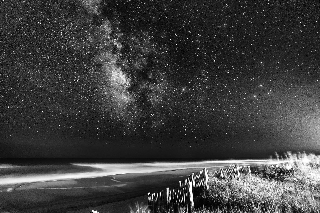 Über einem beleuchteten Strand steigt die Milchstraße auf. Rechts daneben leuchten helle Sterne und Planeten. Das Bild wirkt wie eine Szene aus einem schaurigen Science-Fiction-Film.