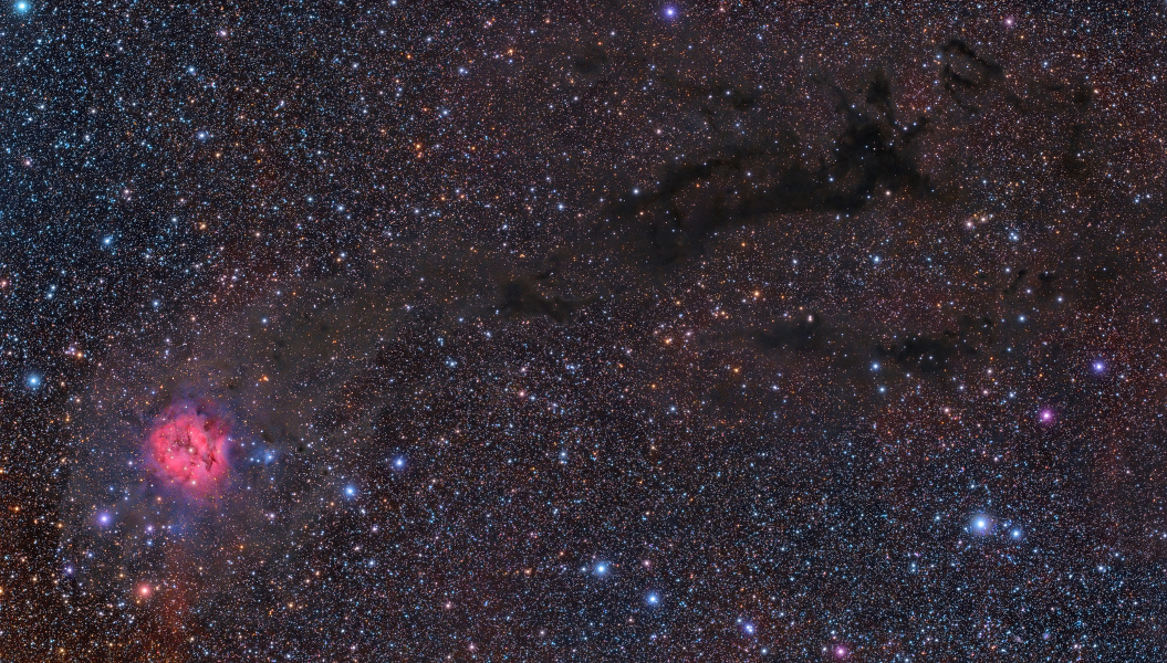 Links unten schimmert ein roter runder Nebel in einer dunklen Himmelsszene, die dicht mit bunten Sternen gesprenkelt ist. Diagonal im Bild verlaufen einige dunkle Staubranken.
