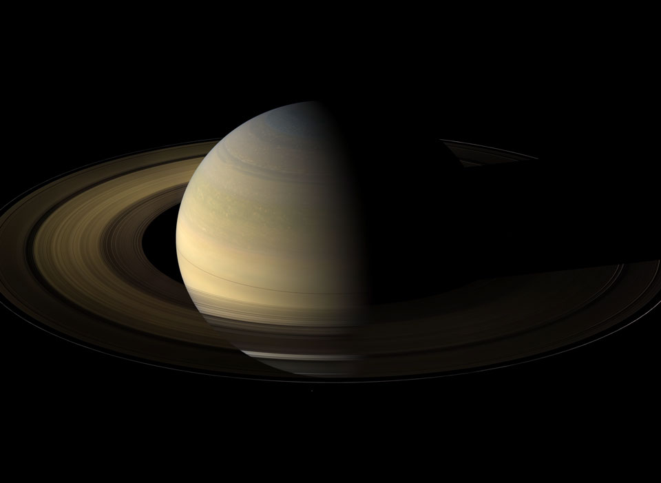 Saturn leuchtet aus dem Dunkel hervor. Links sind die Ringe von der sonnenbeschienen Seite Saturns beleuchtet, rechts sind Planet und Ringe im Dunkel.
