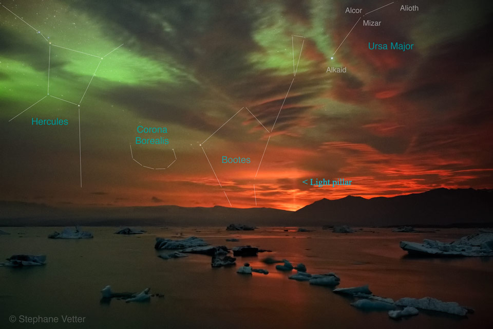 Der wolkige Himmel wirkt dramatisch. Es leuchten Polarlichter, glühende Lava hinter einem Berg und eine Lichtsäule, die ebenfalls vom Vulkanausbruch ausgelöst wird. Vorne ist der Strand eines Gewässers.