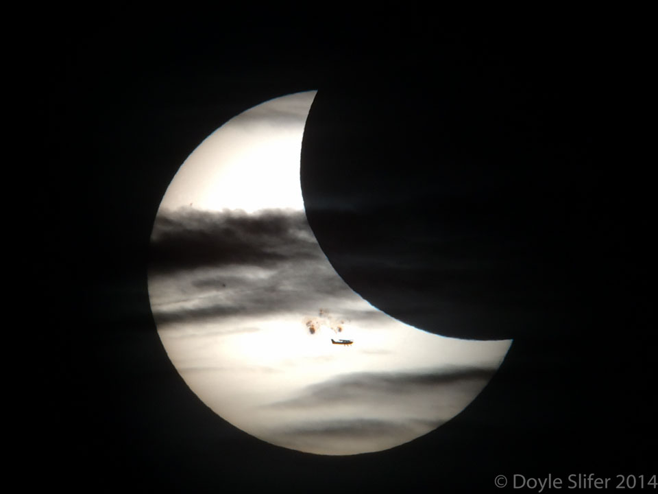 Die Sonne wird nicht nur vom Mond teilweise bedeckt, sondern auch von Wolken, einem Flugzeug und einer Sonnenfleckengruppe. Das Bild zeigt viele Silhouetten und wirkt beinahe wie eine Schwarz-Weiß-Aufnahme.