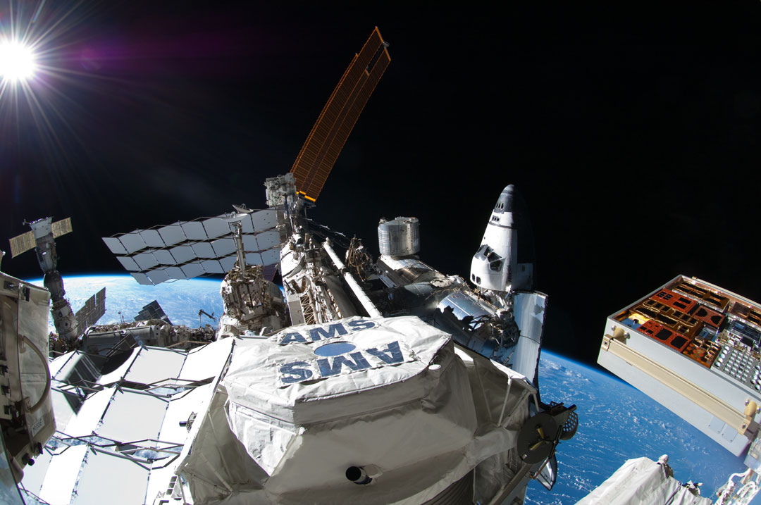 Mitten im Bild ist der AMS-Detektor an Bord der Internationalen Raumstation ISS. Von der Raumstation sind Paneele und Module zu sehen. Rechts ist eine Raumfähre angedockt, dahinter schimmert die blaue Erde. Links oben strahlt die Sonne im schwarzen Weltraum.