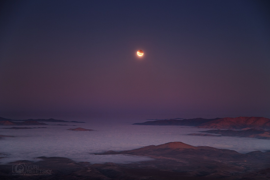 Aus einem Wolkenmeer ragen rötliche Gipfel, darüber ist ein dunkelvioletter Himmel. Mitten im Bild leuchtet der rötliche Vollmond, der etwa halb vom Erdschatten verdunkelt ist. Gerade findet eine Mondfinsternis statt.
