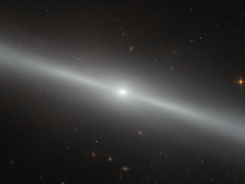 Diagonal verläuft eine helle Linie durchs Bild, sie wirkt verschwommen und hat ein helles Zentrum. Zu welcher Galaxienart sie gehört, sehen wir nicht. Im Hintergrund sind weitere Galaxien verteilt.