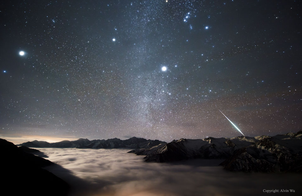Am sternklaren Himmel blitzt rechts unten eine Feuerkugel der Geminiden auf. In den Tälern zwischen den Bergen gleitet ein Nebelmeer. Hinten leuchtet das Sternbild Orion und der helle Stern Sirius.