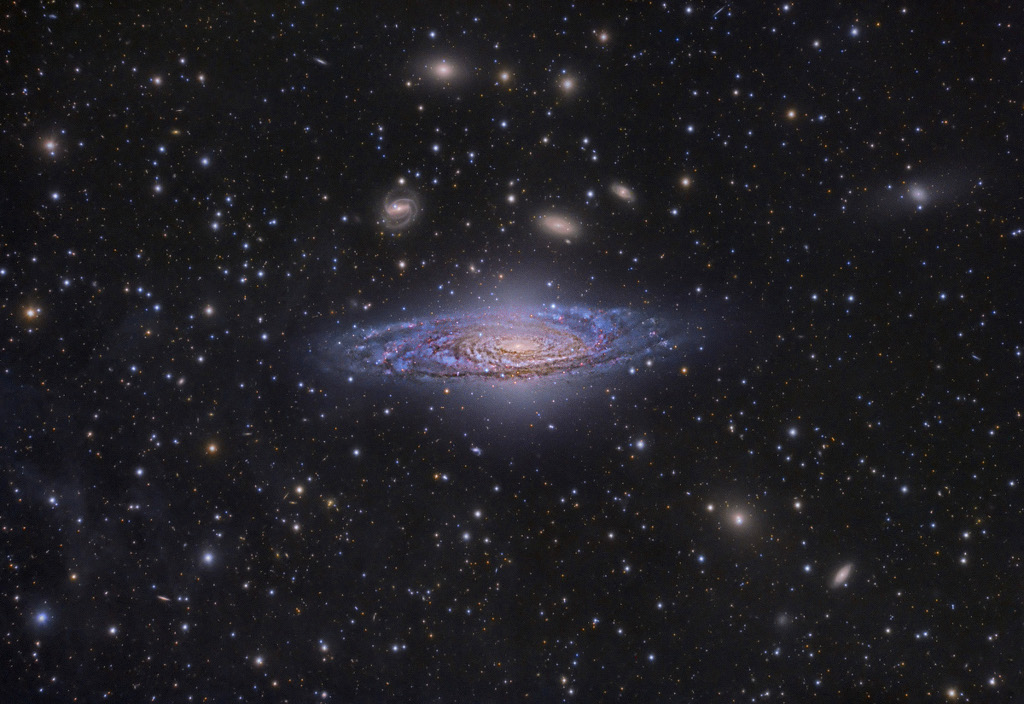 Von Sternen umgeben schwebt eine blaue Spiralgalaxie mit mächtigen dunklen Staubbahnen in den Spiralarmen schräg vor uns. Dahinter sind viele kleinere Galaxien verteilt.