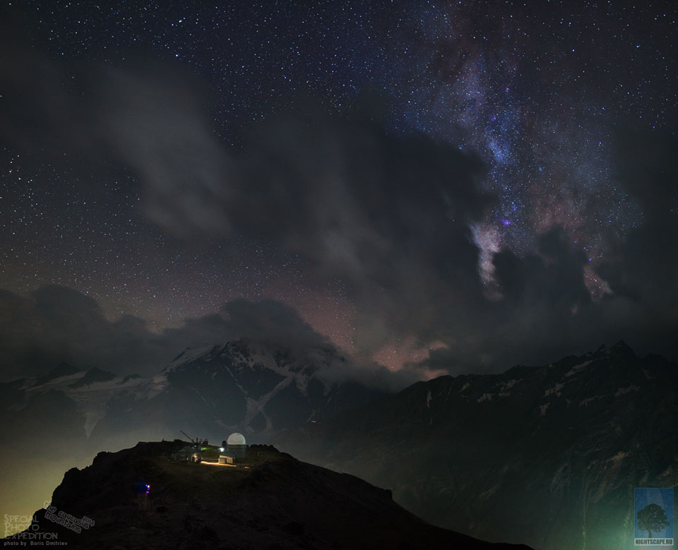 Das Bild zeigt das Pik-Terskol-Observatorium. Es steht im Norden des Kaukasus in Russland. Hinter Wolken und Berggipfeln steigt die Milchstraße auf.