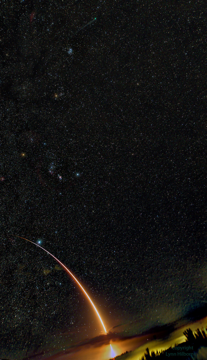 Die leuchtende Feuerspur einer Atlas V zieht am hellen Stern Sirius vorbei. Darüber leuchten das Sternbild Orion, die Hyaden, die Plejaden und der Komet Lovejoy.