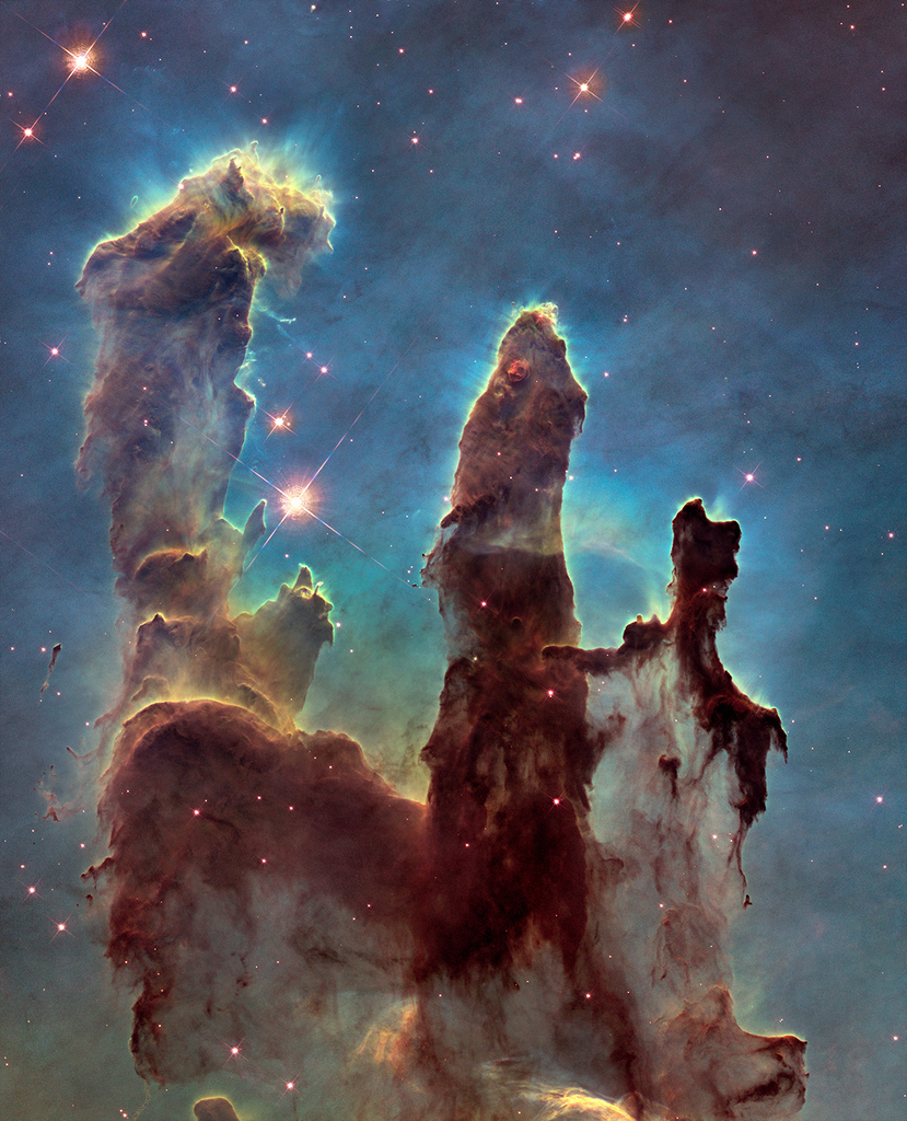 Dunkelbraun ragen die Säulen der Sternbildung im Adlernebel M16 im Sternbild Schlange auf. Dieses Hubble-Bild zeigt sie von einem cyanfarben leuchtenden Schein umgeben, der Nebel im Hintergrund leuchtet zartblau.