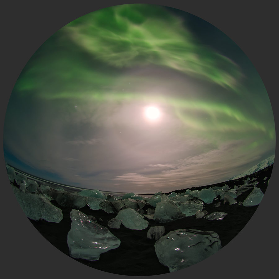 Das Fischaugenbild zeigt unten glitzernde Eisbrocken auf schwarzem Sand. Oben schimmern grüne Polarlichter über dem hellen Mond. Um den Mond verläuft ein 22-Grad-Halo.