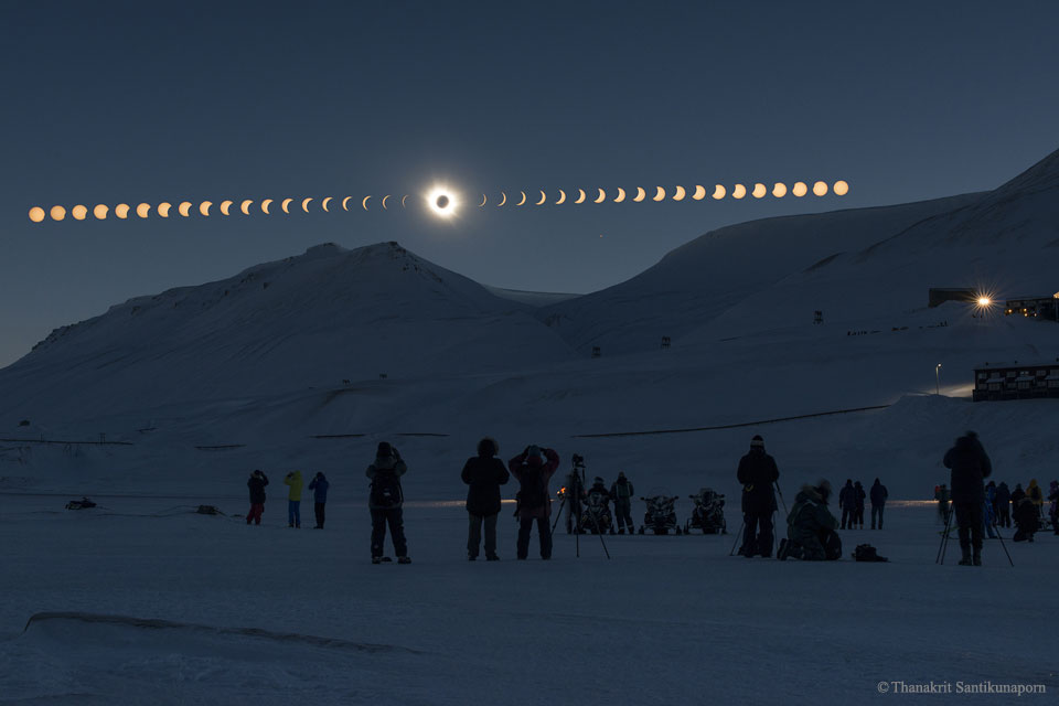 Waagrecht wandern Sonne und Mond über den Himmel. Dabei schiebt sich der Mond immer weiter vor die Sonne, bis er sie in der Mitte bedeckt. Danach gibt er sie langsam wieder frei. Vorne stehen Finsternisjäger im Schatten in einer gebirgigen verschneiten Landschaft.