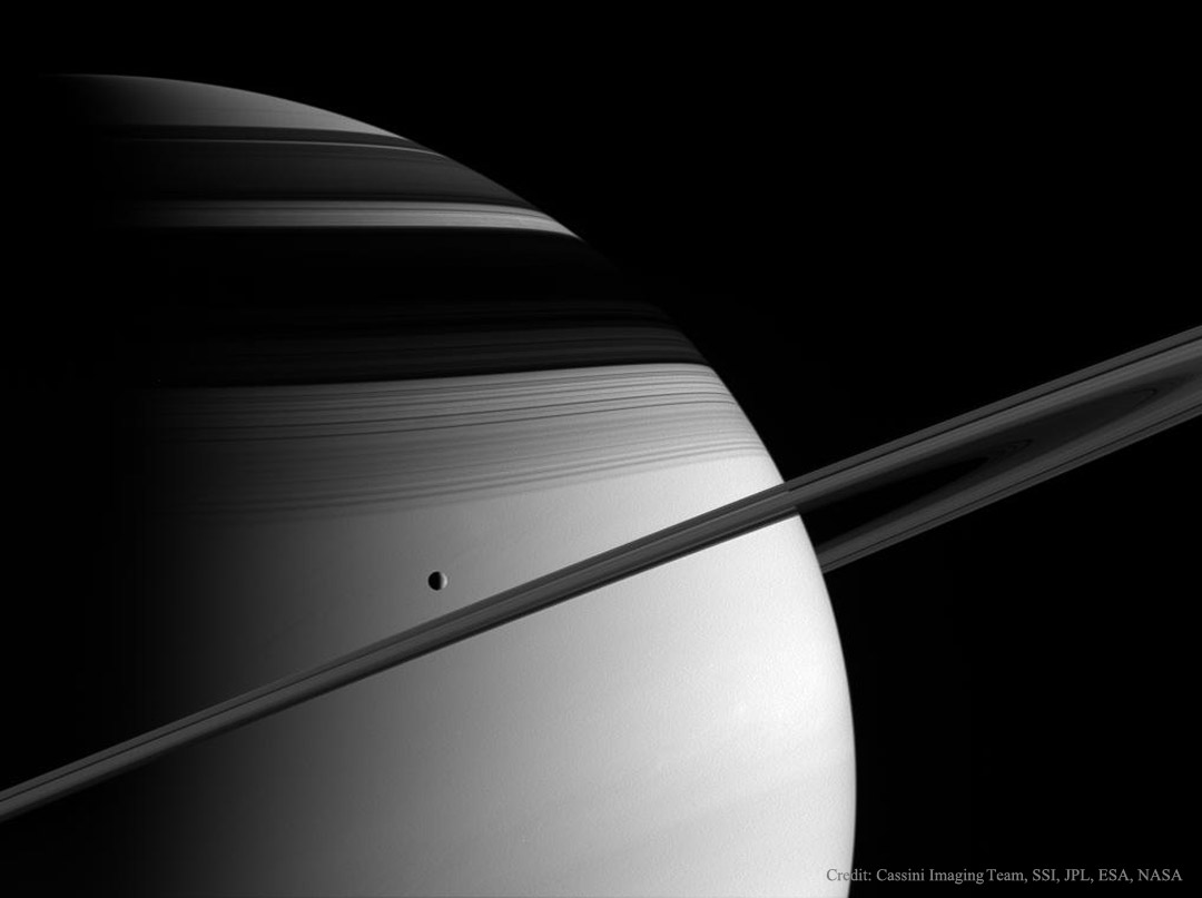 Hinten ragt Saturn riesig auf, die Ringe wirken eher schmal, da wir sie fast von der Kante sehen. Über den Ringen schwebt der Saturnmond Tethys im Sonnenlicht. Die Ringe werfen einen breiten Schatten auf den Planeten Saturn.