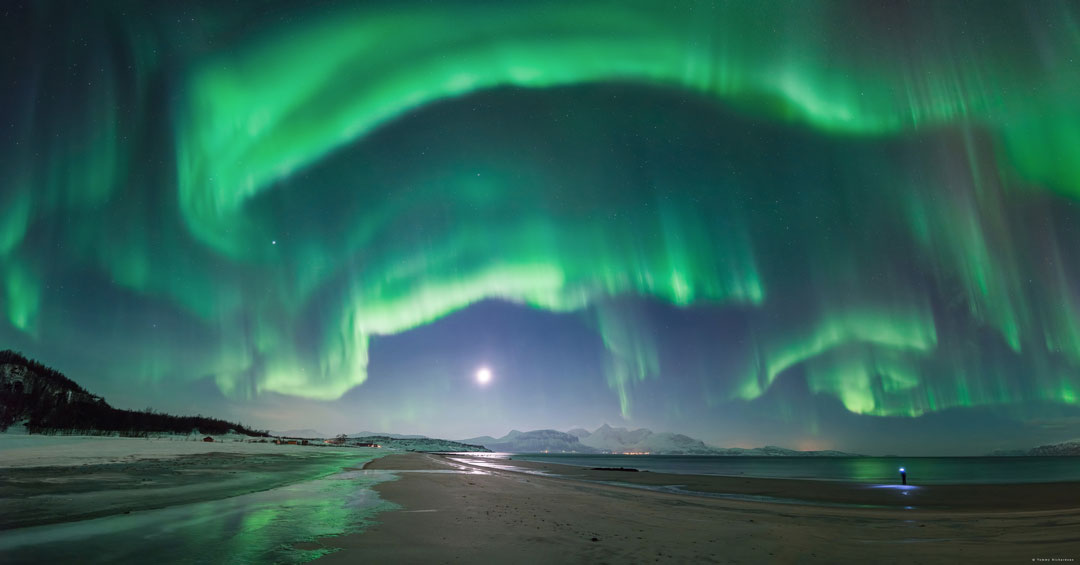 Dichte Polarlichtschleier hängen vom Himmel, sie schimmern grün. Unten leuchtet der Mond über dem Strand in Norwegen. Rechts sucht eine Person mit Taschenlampe einen Objektivdeckel.