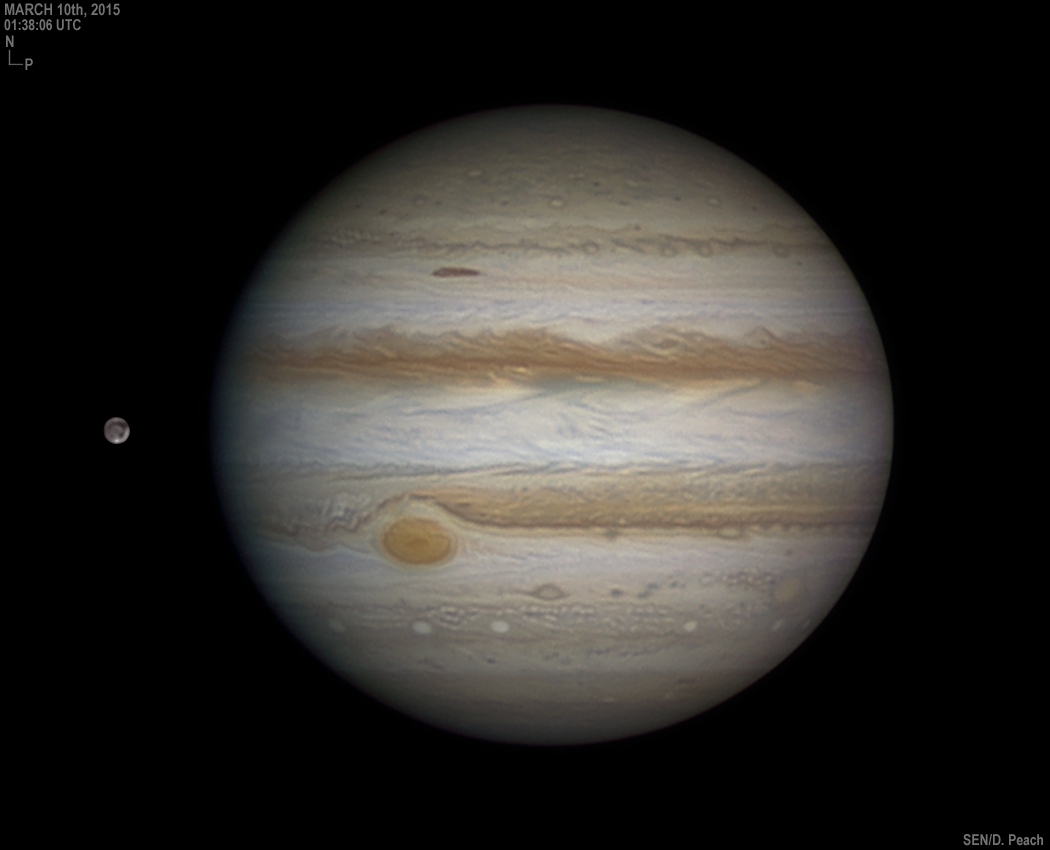 Links neben dem voll beleuchteten Jupiter schwebt der Mond Ganymed. Auf Jupiter zeichnen sich zwei markante braune Wolkenbänder zwischen beigen Wolken ab. In einem davon befindet sich der Rote Fleck links neben der Mitte.