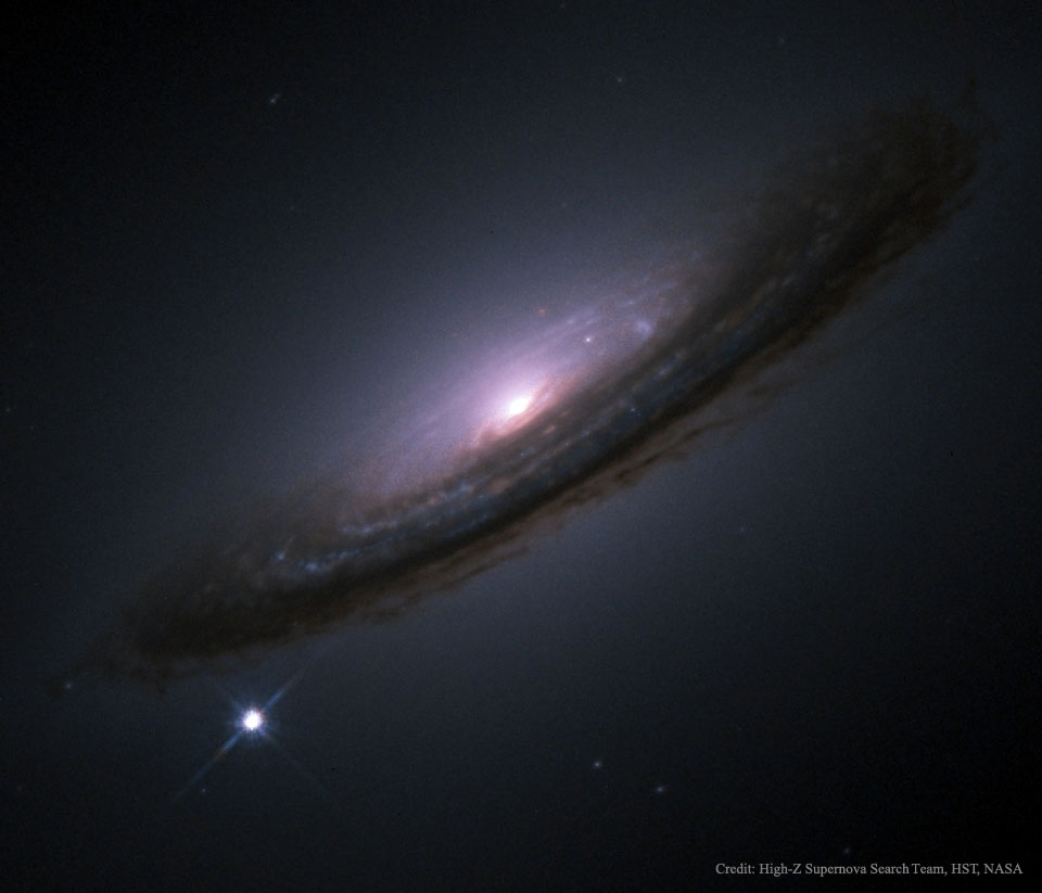Schräg im Bild ist eine düstere Galaxie. Ihr Kern ist hier violett-rosa abgebildet, nur das Zentrum leuchtet hell. Am Rand verlaufen dicke Staubbahnen. Links unten strahlt eine helle Supernova.