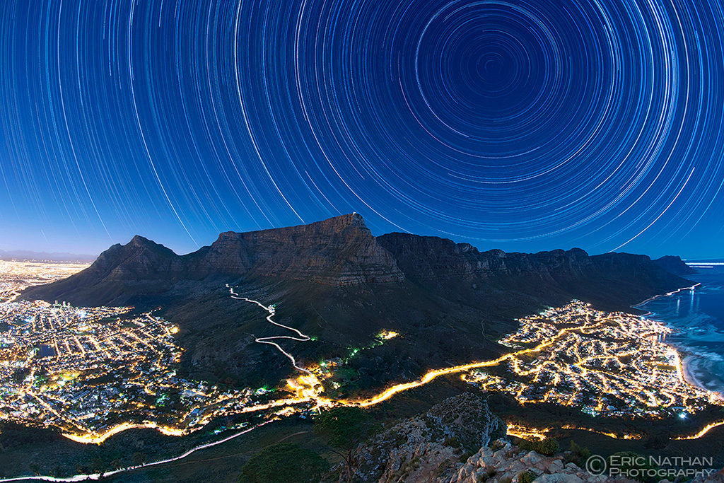 Über Kapstadt ziehen Sterne am blauen nächtlichen Himmel ihre kreisförmigen Strichspuren. Der Tafelberg ragt aus den hellen Lichtern der Stadt heraus.