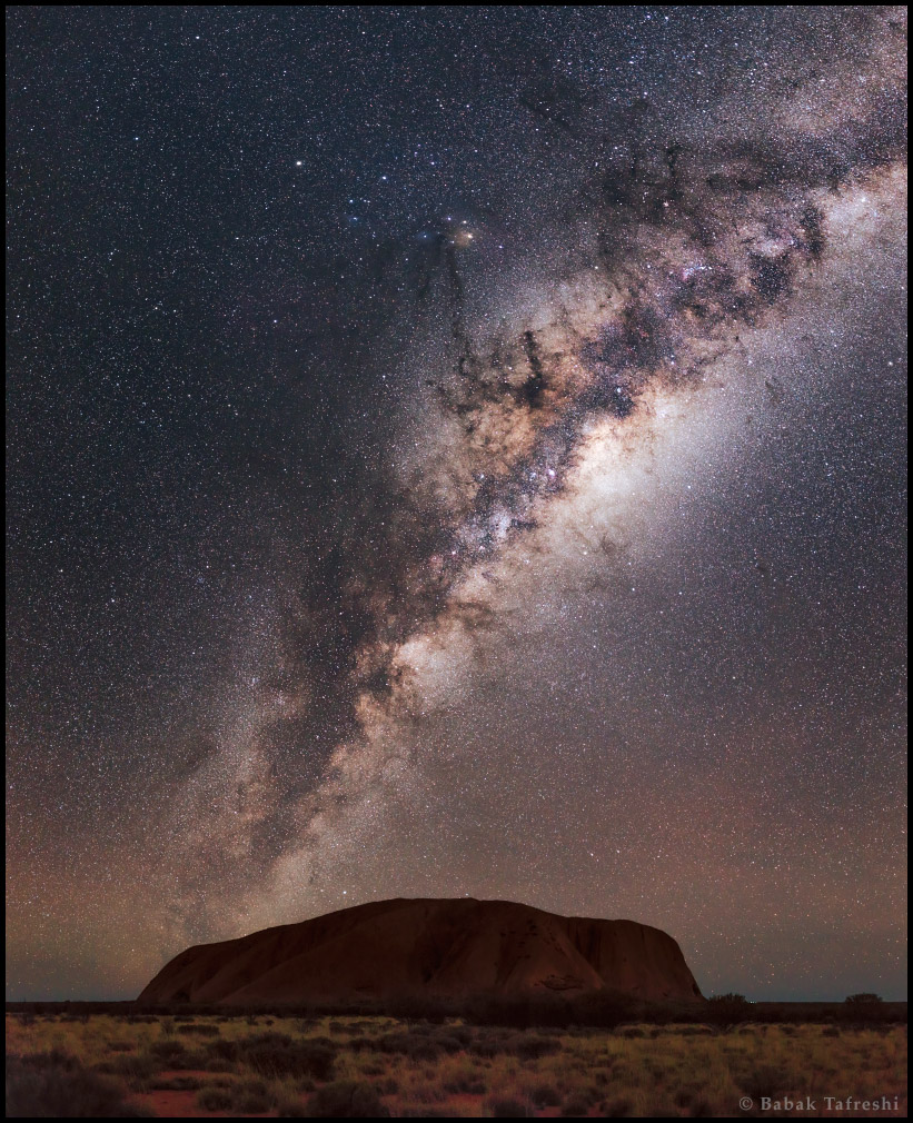 Aus einer weiten Ebene ragt der Uluru hoch, er ist ein australisches Wahrzeichen. Darüber steigt schräg die Milchstraße auf, sie ist von markanten dunklen Staubwolken strukturiert.
