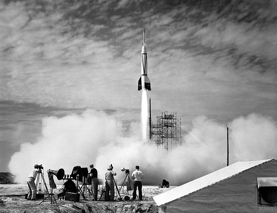 Eine Rakete startet und stößt unten einen weißen Strahl aus. Vorne wird das Ereignis gefilmt und fotografiert.