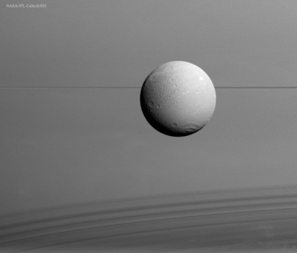 Hinter dem Saturnmond Dione verlaufen die Saturnringe als dünner, dunkler Strich. Darunter fallen die Schatten der Ringe auf die Oberfläche von Saturn und bilden ein wellenartiges Muster.