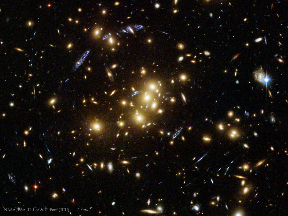 Mitten im Bild ist eine Ansammlung ellliptischer, gelb leuchtender Galaxien. Außen herum sind blaue Bögen angeordnet, die wohl alle zu derselben Galaxie im Hintergrund gehören. Das ganze Bild ist voller Galaxien.