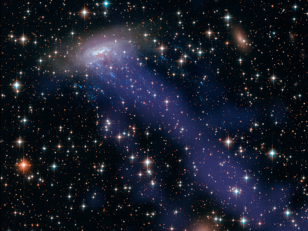 Eine Spiralgalaxie rast unglaublich schnell diagonal durchs Bild nach rechts oben. Sie zieht einen langen, blau leuchtenden Strahl aus abgestreiftem Gas und Staub hinter sich her.