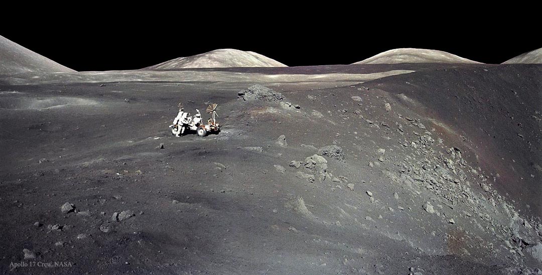 Am Rand eines Kraterwalls, der nach rechts abfällt, steht ein Astronaut mit einem Mondrover. Dahinter ist ebenes Gelände, hinter dem einige runde Berge aufragen. Der Himmel oben ist schwarz.