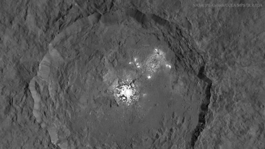 Mitten im Bild ist ein Krater, er ist unten nicht ganz dargestellt. In der Mitte ist ein großer, ausgefranster heller Fleck, links oben sind weitere verstreute und verschwommene Flecken.