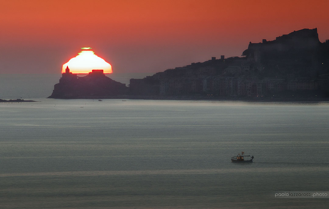 Hinter dem Meer geht die Sonne über der Kirche San Pietro im italienischen Porto Venere unter. Sie wirkt eigentümlich geschichtet. Die oberste Schicht hat einen grünen Rand. Der Himmel dahinter leuchtet rot.