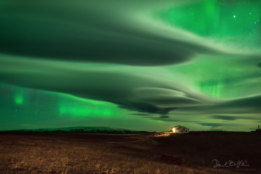 Am Horizont steht ein Haus, an dem ein Licht leuchtet. Darüber ziehen Wolken über den Himmel. Durch die Lücken schimmert grünes Polarlicht.