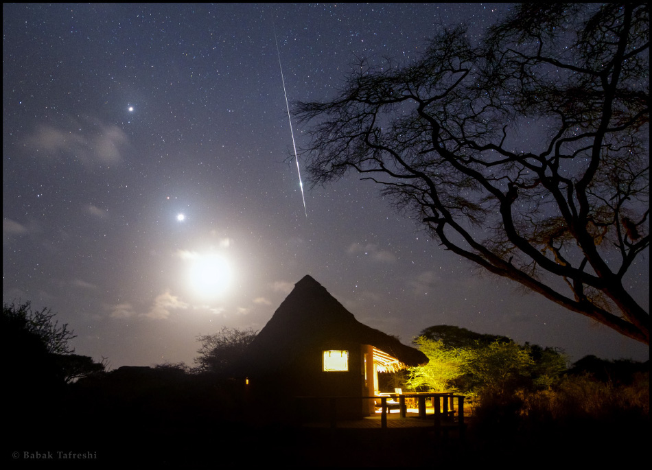 Über einer Hütte, die von innen beleuchtet ist, fällt eine Feuerkugel vom Himmel. Links stehen der helle Mond, Venus, Mars und Jupiter teil übereinander in einer Reihe am Himmel.
